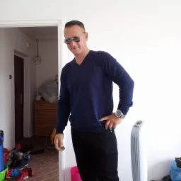 Mężczyzna w okularach pozuje w pomieszczeniu z białymi ścianami, ubrany w ciemny sweter i czarne spodnie. W tle widoczny wentylator i otwarte drzwi.