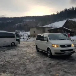 Biały bus Volkswagen i autokar na tle zimowego krajobrazu z budynkami i zalesionym wzgórzem.