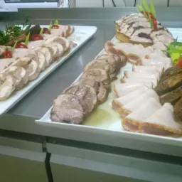 Dwa białe prostokątne półmiski z różnorodnymi wędlinami i dekoracjami z ziół i papryki, ustawione na metalowym blacie kuchennym.