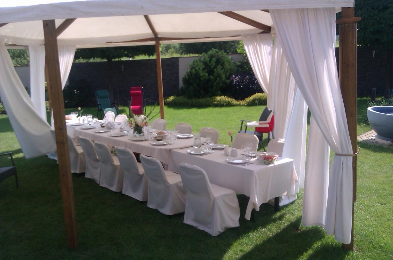 Uroczysty stół nakryty białym obrusem i zastawą, otoczony białymi krzesłami w ogrodzie pod baldachimem z białymi zasłonami.