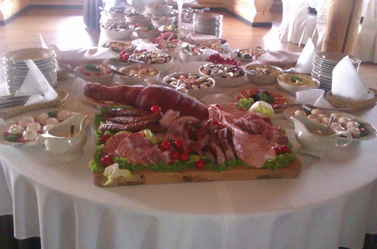 Stół bufetowy z różnorodnymi daniami mięsnymi, wędlinami, sałatkami i talerzami, przygotowany na uroczystość, z widoczną pieczenią, kiełbasami i dekoracją z pomidorków cherry.