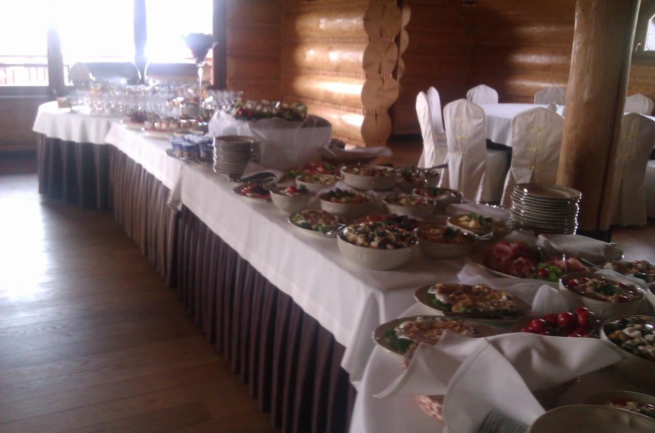 Stół bufetowy z różnorodnymi sałatkami, przekąskami i deserami, ustawiony w rustykalnym wnętrzu z drewnianymi ścianami i kolumnami, nakryty białym obrusem z brązową falbaną.