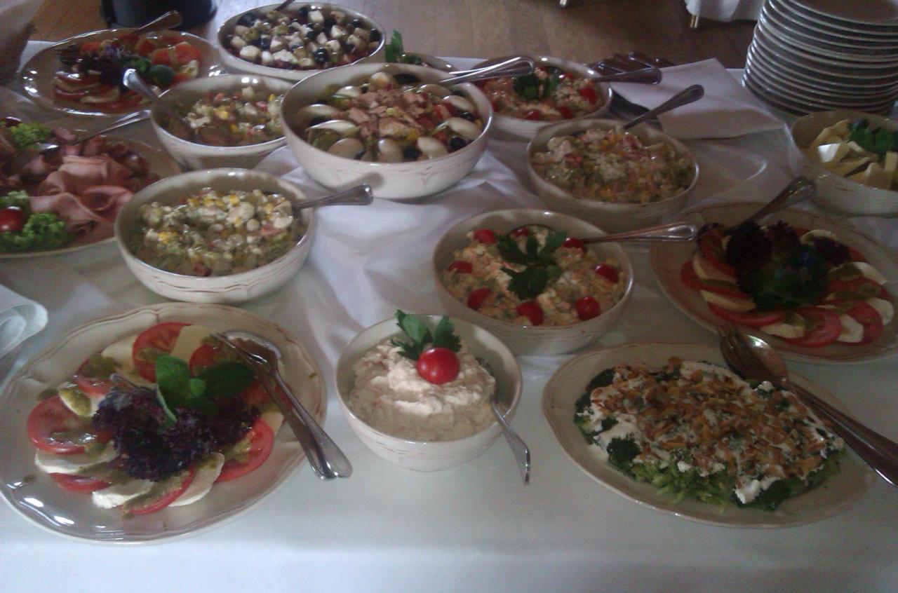 Stół z białym obrusem zastawiony różnorodnymi sałatkami w misach i na talerzach, widoczne sztućce, kompozycje z pomidorów, mozzarelli i zieleniny, sałatki warzywne i z dodatkiem mięs.