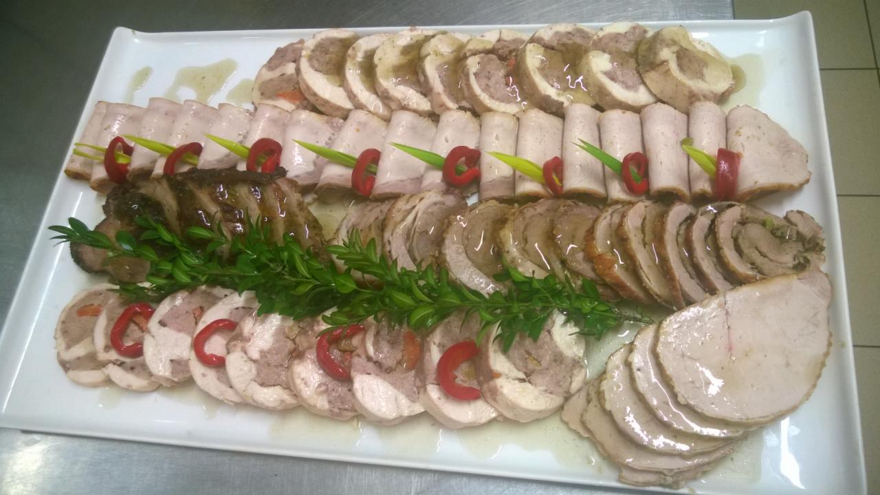 Dekoracyjnie ułożony półmisek wędlin i mięs w galarecie, ozdobiony gałązkami zieleni i papryką, na białym talerzu.