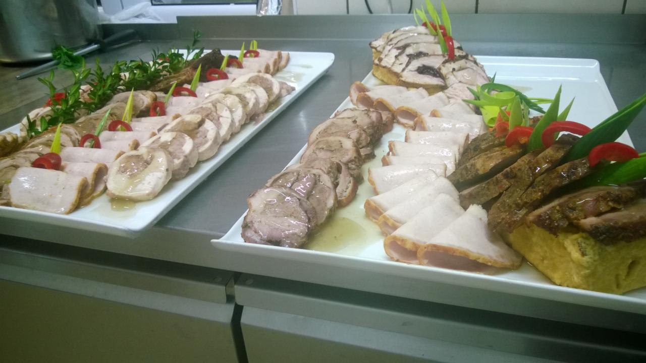 Dwa białe prostokątne półmiski z różnorodnymi wędlinami i dekoracjami z ziół i papryki, ustawione na metalowym blacie kuchennym.