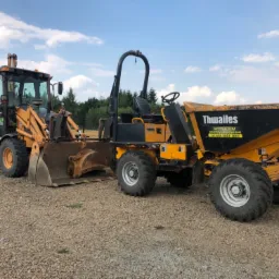 Żółto-czarna koparko-ładowarka CASE 580 Super L i wywrotka Thwaites na żwirowym terenie, otoczone zielenią drzew, w tle błękitne niebo z chmurami.