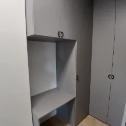 Szara zabudowa meblowa z biurkiem wnękowym i szafami z minimalistycznymi uchwytami. Jasna podłoga, stonowana kolorystyka, nowoczesny design.