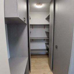 Sylwestri Usługi remontowo-budowlane - Wnętrze garderoby z szarymi szafkami, półkami i oświetleniem sufitowym. Widok z perspektywy wejścia, ukazujący minimalistyczny design i funkcjonalne rozwiązanie przechowywania.