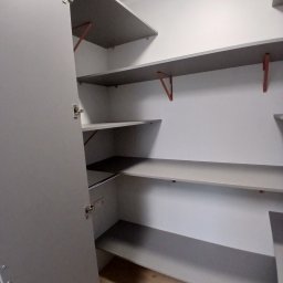 Sylwestri Usługi remontowo-budowlane - Wnętrze szafy wnękowej z szarymi półkami i drzwiami. Widoczne metalowe zawiasy i wsporniki półek. Podłoga z paneli. Minimalistyczny design.