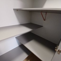 Sylwestri Usługi remontowo-budowlane - Wnętrze szafy z szarymi półkami i metalowymi wspornikami w kolorze pomarańczowym. Widoczne zawiasy drzwi, jasna podłoga. Ujęcie z dołu, perspektywa wnętrza.