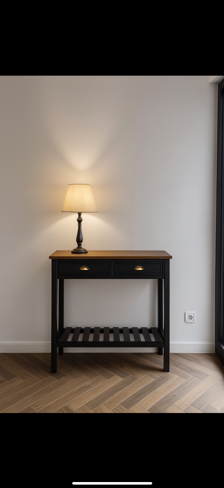 Stylowa konsola z dwoma szufladami i półką, oświetlona lampą stołową, ustawiona na parkiecie w jodełkę, obok okna. Minimalistyczne wnętrze.