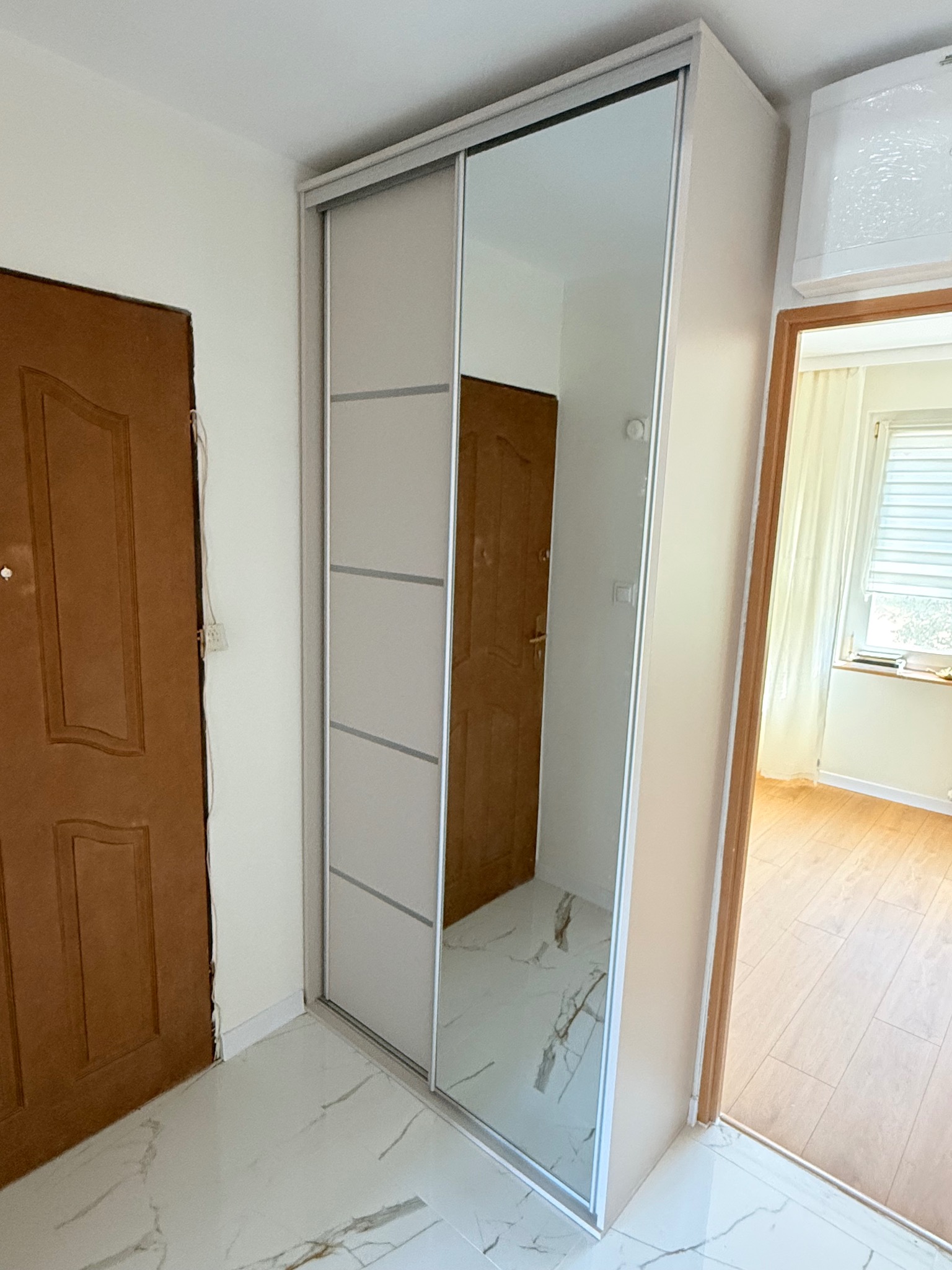 Jasna szafa przesuwna z lustrem w przedpokoju, widok na wnętrze mieszkania. Drzwi wejściowe po lewej, marmurowe płytki podłogowe, białe ściany.