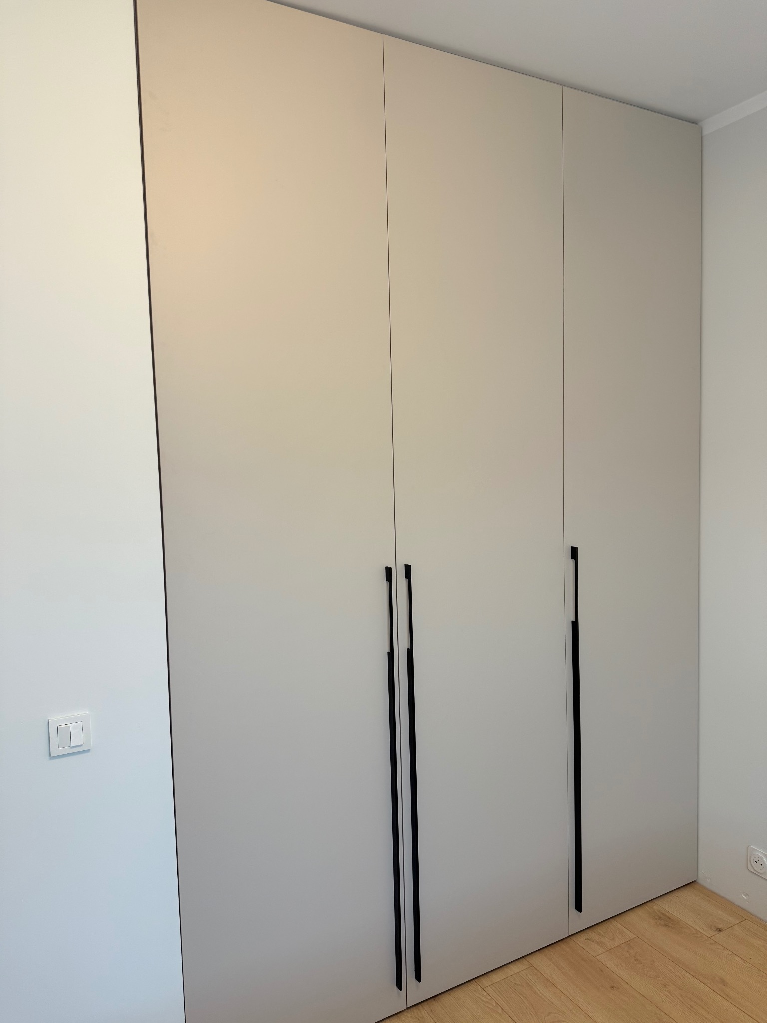 Nowoczesna, beżowa szafa wnękowa z długimi, czarnymi uchwytami, idealnie dopasowana do jasnego wnętrza z drewnianą podłogą i gniazdkiem elektrycznym przy podłodze.