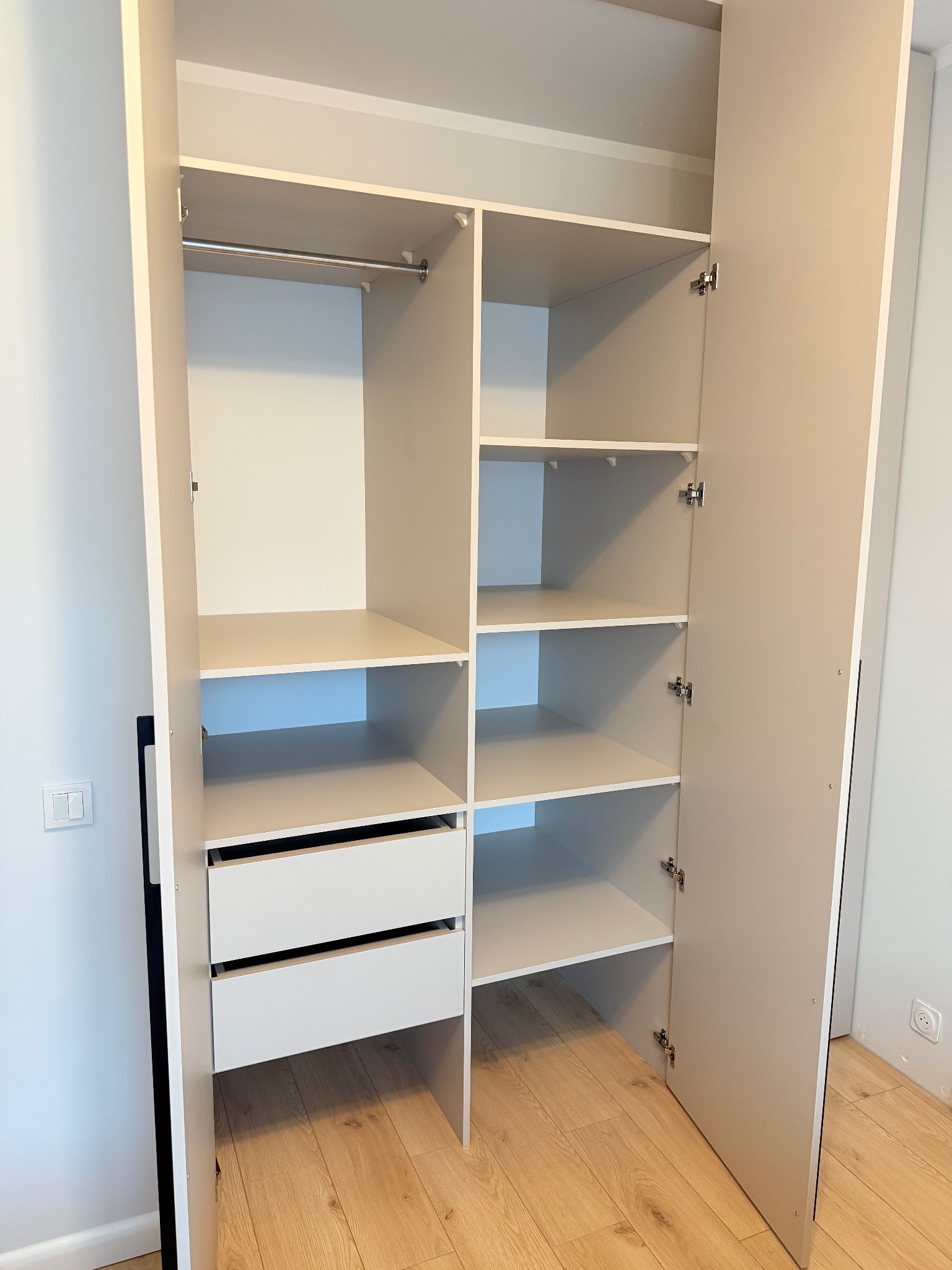 Jasna szafa z otwartymi drzwiami, widoczne półki, drążek i dwie szuflady. Minimalistyczny design, drewniana podłoga, gniazdko elektryczne w tle.