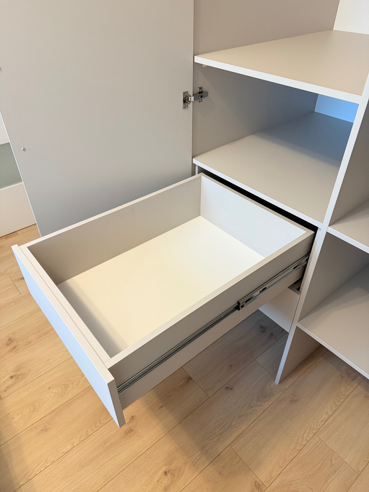 Jasnoszara szafa na wymiar z otwartą szufladą na prowadnicach, widoczne półki i zawiasy drzwi. Parkiet z jasnego drewna w tle.