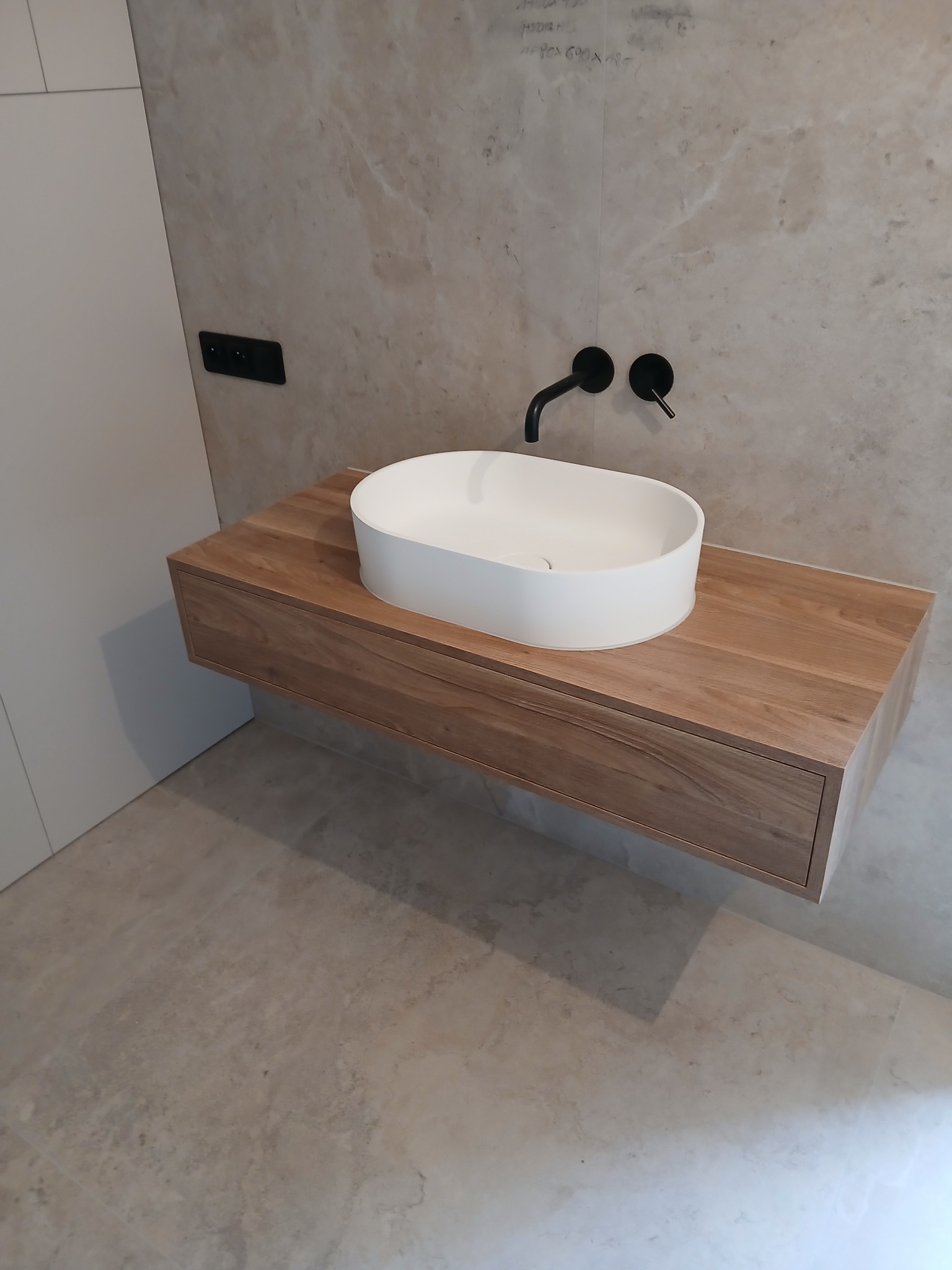 Nowoczesna łazienka: biała, owalna umywalka nablatowa z czarną armaturą na szafce z drewna o ciepłym odcieniu, na tle beżowej ściany i podłogi z płytek.