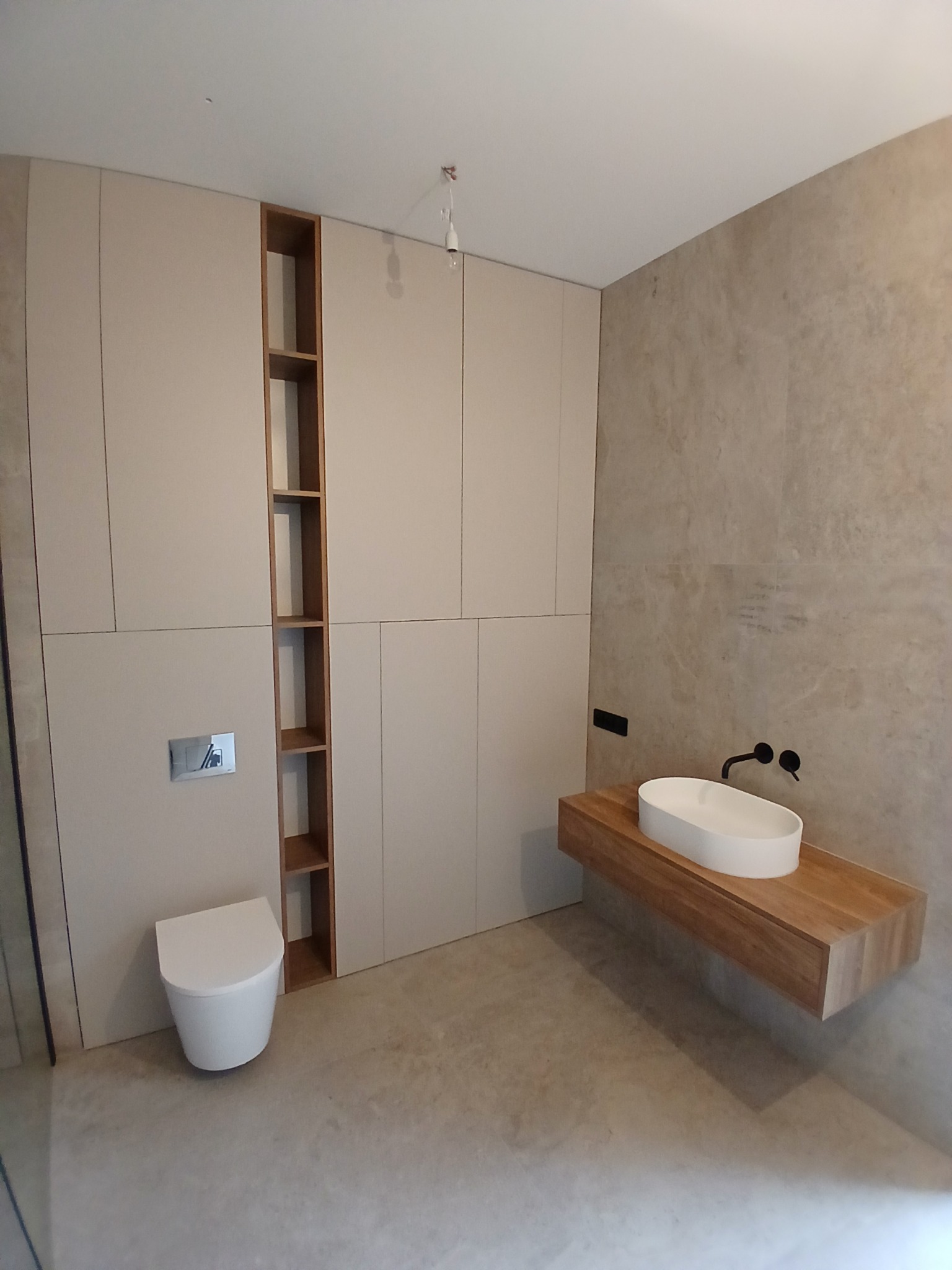 Nowoczesna łazienka z zabudową meblową w beżach, drewnianym blatem pod umywalkę i wiszącą toaletą. Minimalistyczny design, neutralna kolorystyka, kamienne płytki na ścianie.