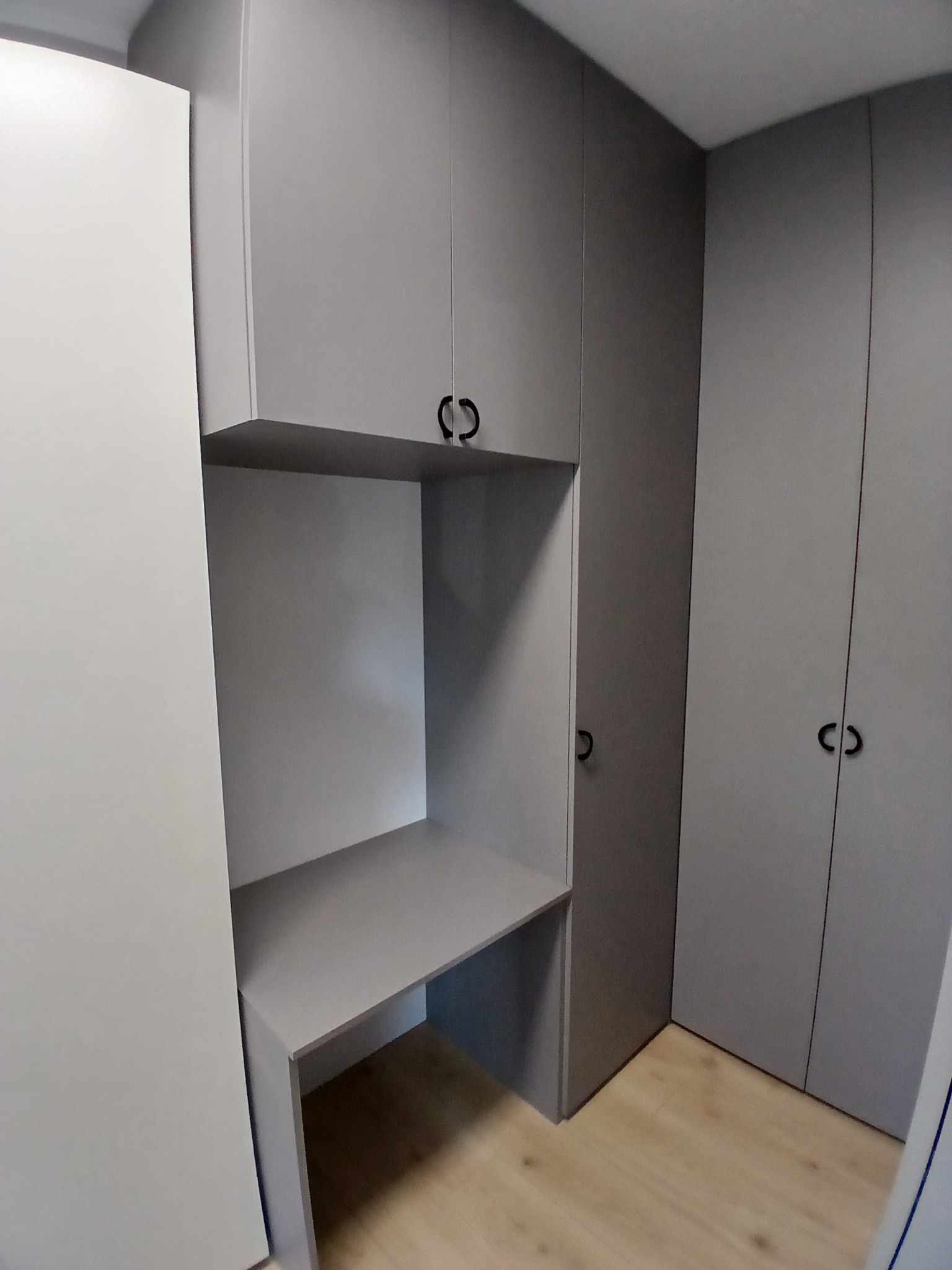 Szara zabudowa meblowa z biurkiem wnękowym i szafami z minimalistycznymi uchwytami. Jasna podłoga, stonowana kolorystyka, nowoczesny design.