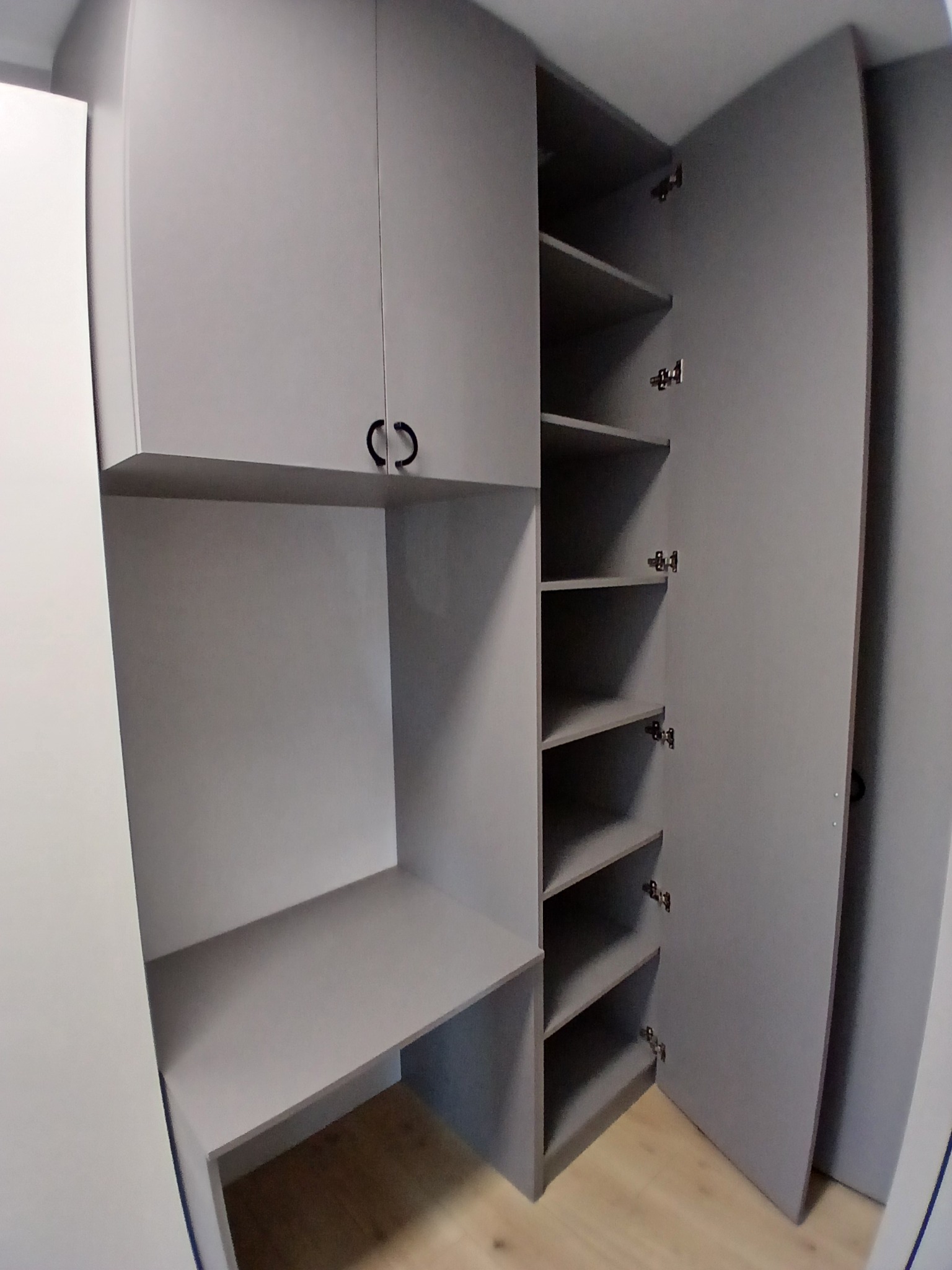 Szara szafa wnękowa z otwartymi drzwiami i półkami, zintegrowana z biurkiem. Minimalistyczny design, widoczne okucia i drewniana podłoga.