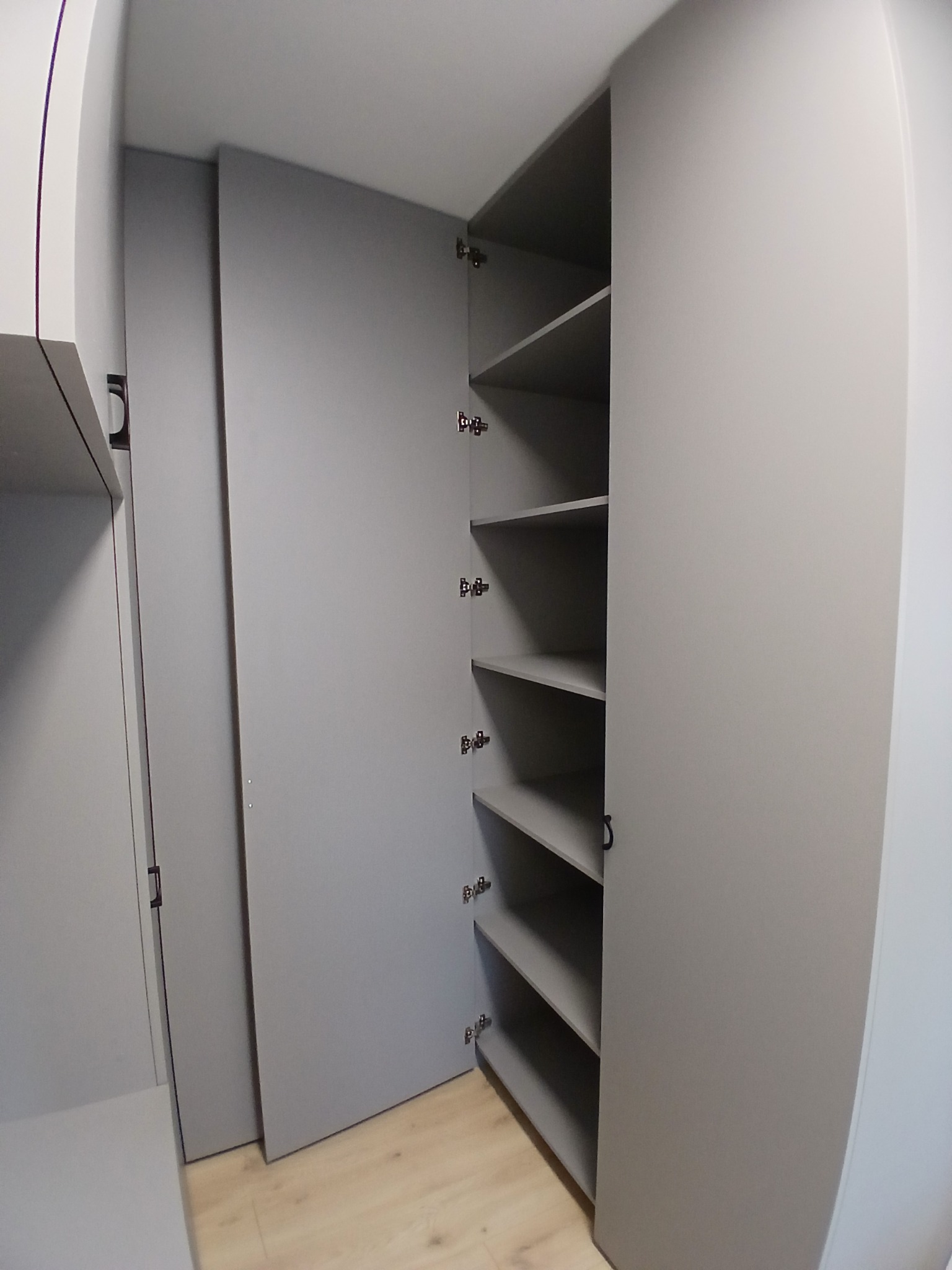 Szara szafa na wymiar z otwartymi drzwiami, ukazująca puste półki. Widoczny fragment podłogi z paneli. Minimalistyczny design, neutralna kolorystyka, precyzyjne wykonanie.