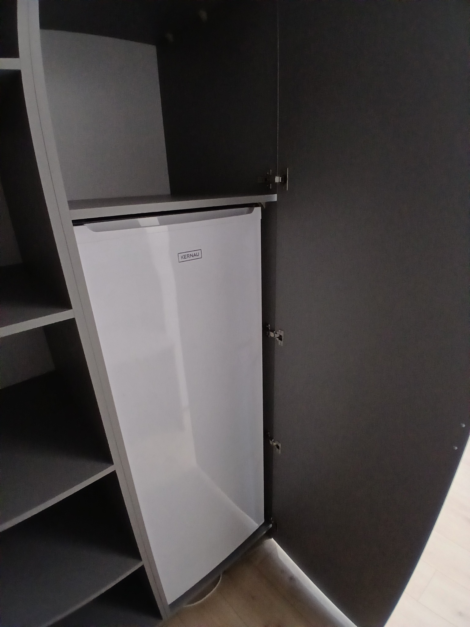 Szara szafa na wymiar z półkami i wbudowaną białą lodówką marki Kernau. Otwarta szafka prezentuje funkcjonalne rozwiązanie do przechowywania.