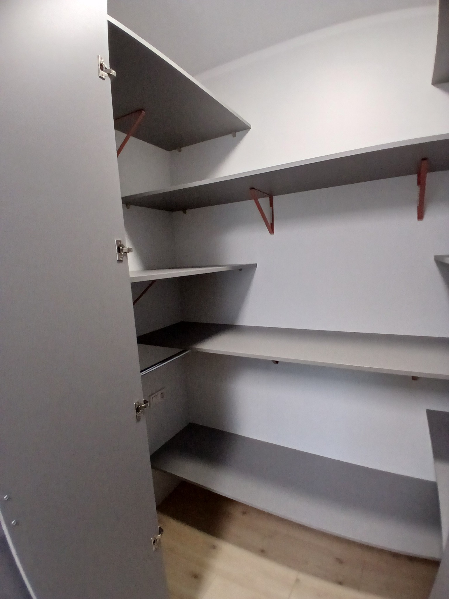 Wnętrze szafy wnękowej z szarymi półkami i drzwiami. Widoczne metalowe zawiasy i wsporniki półek. Podłoga z paneli. Minimalistyczny design.