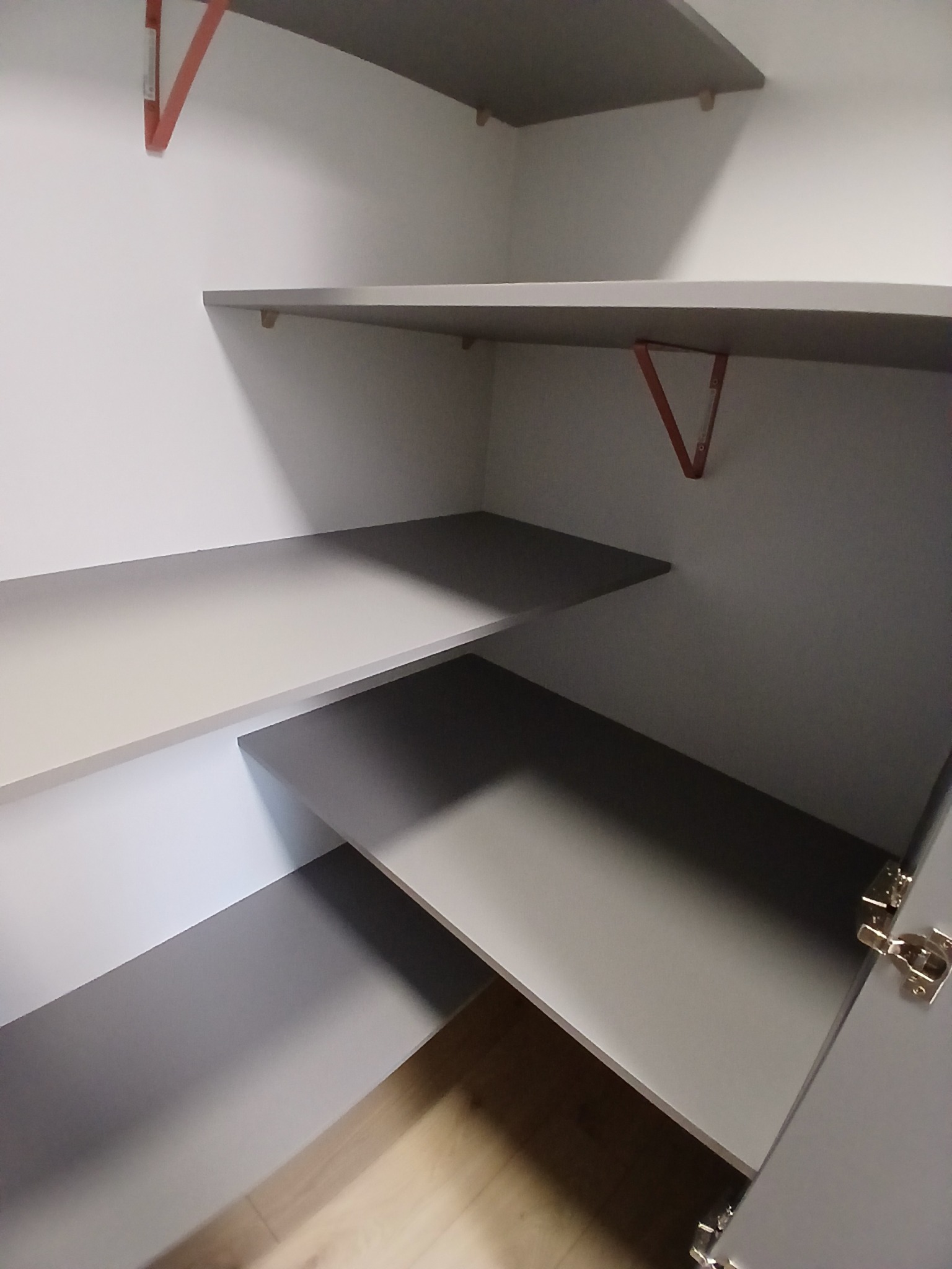 Wnętrze szafy z szarymi półkami i metalowymi wspornikami w kolorze pomarańczowym. Widoczne zawiasy drzwi, jasna podłoga. Ujęcie z dołu, perspektywa wnętrza.