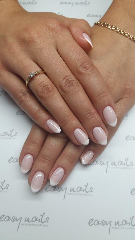 Dłonie kobiety z delikatnym french manicure na owalnych paznokciach, złoty pierścionek z brylantem i bransoletka na tle firmowego podkładu.