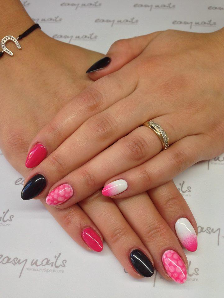 Dłonie kobiety z manicure hybrydowym w kolorach różu, bieli i czerni, z różnymi wzorami, w tym ombre i różowe kropki, na tle z delikatnym wzorem logo salonu, z bransoletką z podkową i złotym...