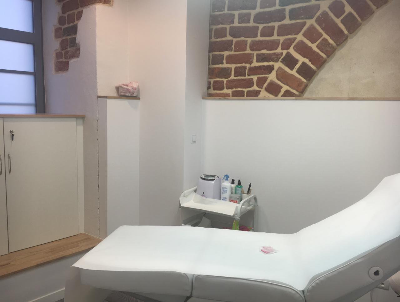 Jasne wnętrze gabinetu kosmetycznego z leżanką pokrytą białym prześcieradłem, w tle widoczny wózek z akcesoriami i fragment ściany z dekoracyjnym motywem cegieł.