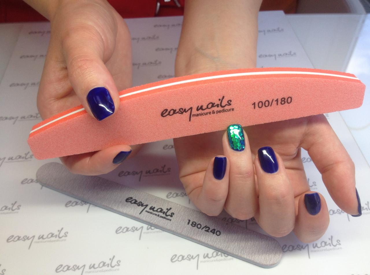Dłonie z pomalowanymi paznokciami w odcieniach granatu i zieleni, trzymające pilniki do paznokci o różnej gradacji (100/180 i 180/240) z logo 'easy nails manicure & pedicure'.