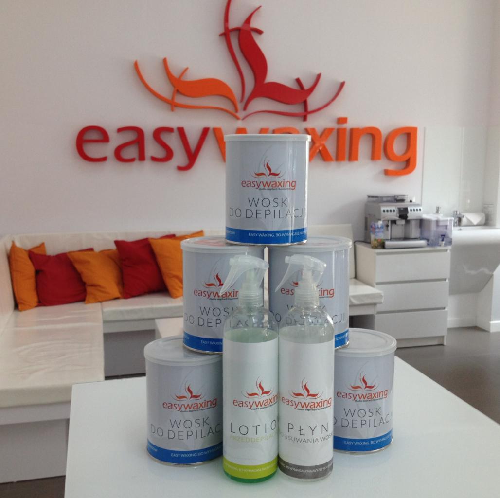 Piramida z puszek wosku do depilacji EasyWaxing i butelek z płynami przed i po depilacji, na tle sofy z pomarańczowymi poduszkami i logiem firmy na ścianie.