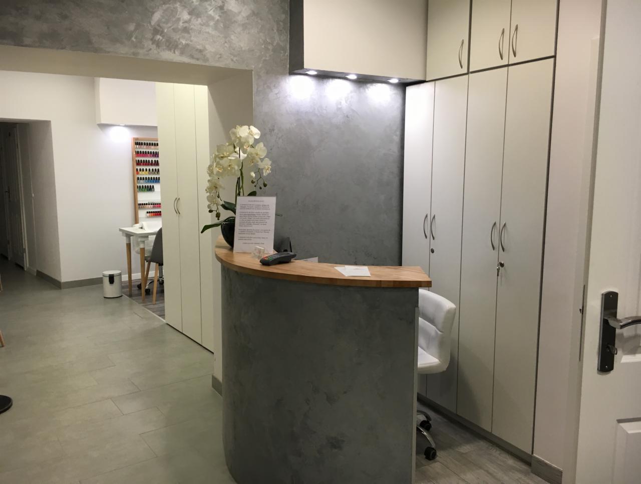 Recepcja w salonie kosmetycznym z widocznym stanowiskiem do zabiegów na paznokcie w tle, półokrągły blat z drewna, dekoracja kwiatowa i szara ściana z efektem betonu.