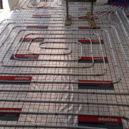 Ułożone rury ogrzewania podłogowego na folii z siatką, widoczne złącza i fragmenty instalacji hydraulicznej w tle.