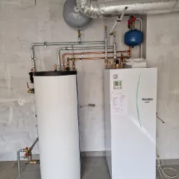 Biały zasobnik ciepłej wody i pompa ciepła Ecodan połączone rurami miedzianymi i stalowymi na tle surowej ściany z instalacją wentylacyjną pod sufitem.