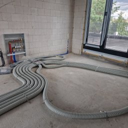 Eco Instal Plus, Robert Kolbus - Szereg szarych rur karbowanych instalacji grzewczej, ułożonych na surowej betonowej posadzce w pomieszczeniu z rozdzielaczem na ścianie z bloczków i dużym oknem balkonowym.