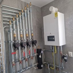 Eco Instal Plus, Robert Kolbus - Kompletna instalacja grzewcza z białym kotłem De Dietrich, rozdzielaczami i pompami obiegowymi, precyzyjnie zamontowana na nowoczesnej, szarej ścianie z płytek.
