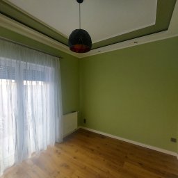 OLIKO Sp. z o.o. - Pokój po malowaniu w Bydgoszczy: zielone ściany, drewniana podłoga, biały sufit z dekoracyjną listwą i nowoczesna lampa wisząca. Jasne zasłony w oknie.