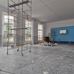 OLIKO Sp. z o.o. - Malowanie sali gimnastycznej w trakcie prac. Widoczne rusztowania, zabezpieczona podłoga folią, puszki z farbą i malarz przy ścianie z koszem.