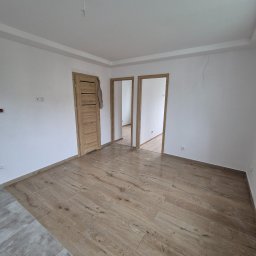 OLIKO Sp. z o.o. - Jasne wnętrze po remoncie: drewnopodobna podłoga, białe ściany, dwoje drzwi z drewnianymi ościeżnicami. Widoczne niedociągnięcia w postaci wystających kabli.