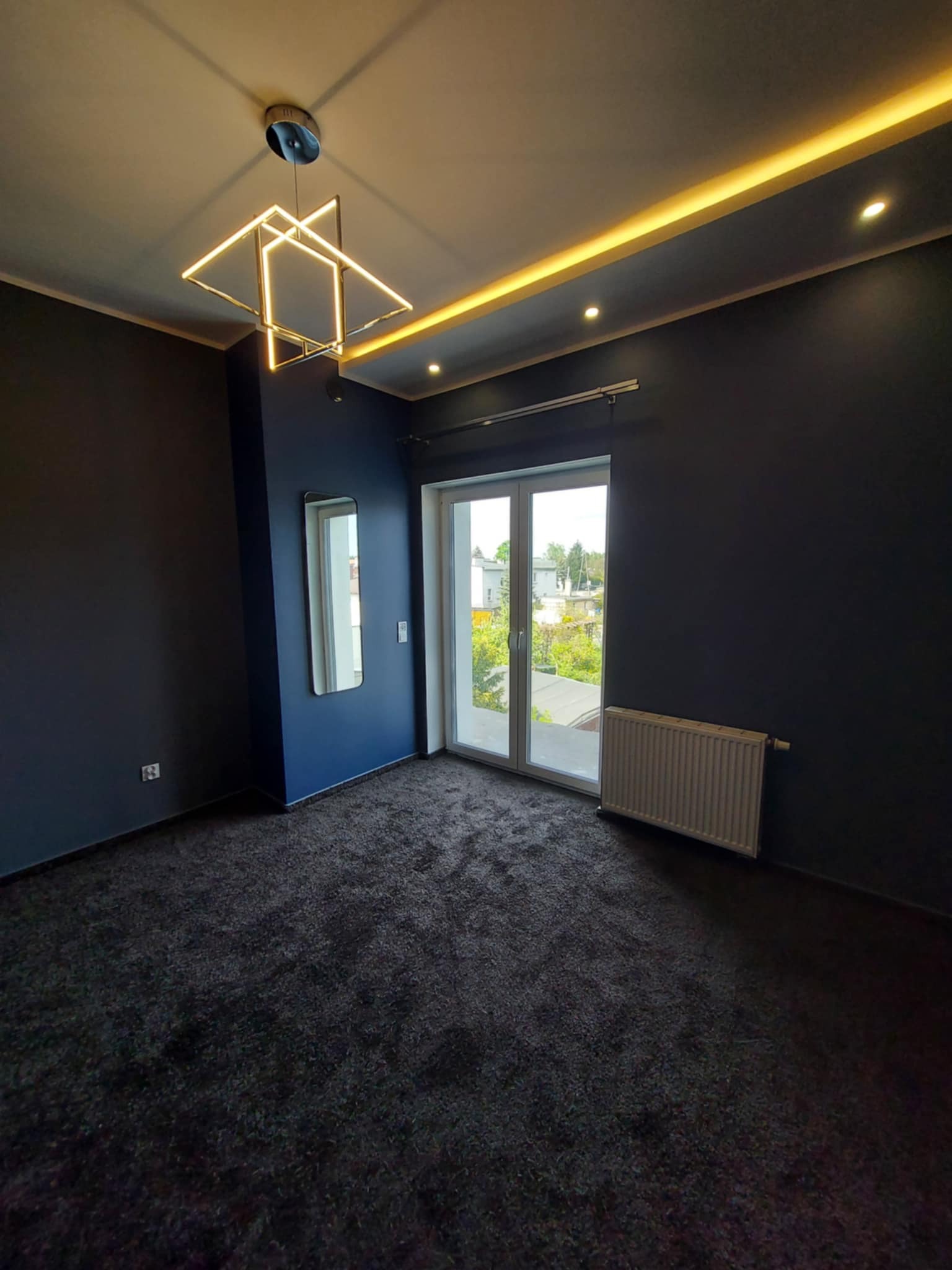 Pokój po remoncie: ciemne ściany, szara wykładzina, nowoczesna lampa sufitowa i okno balkonowe z widokiem na zieleń. Minimalistyczne wnętrze z grzejnikiem.