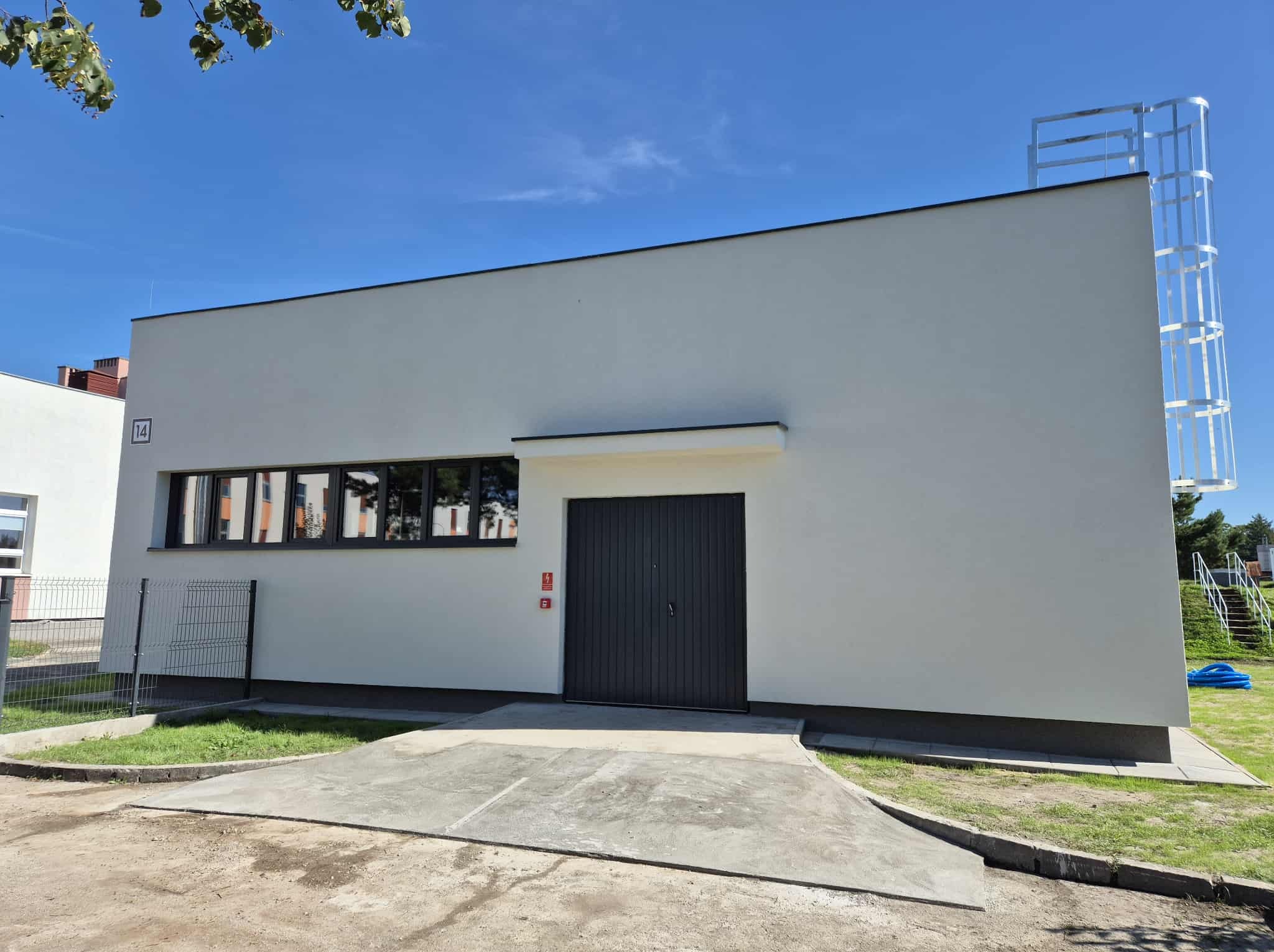 Biały, modernistyczny budynek z ciemnymi oknami i drzwiami garażowymi, widoczny z zewnątrz w słoneczny dzień. Na dachu metalowa konstrukcja.