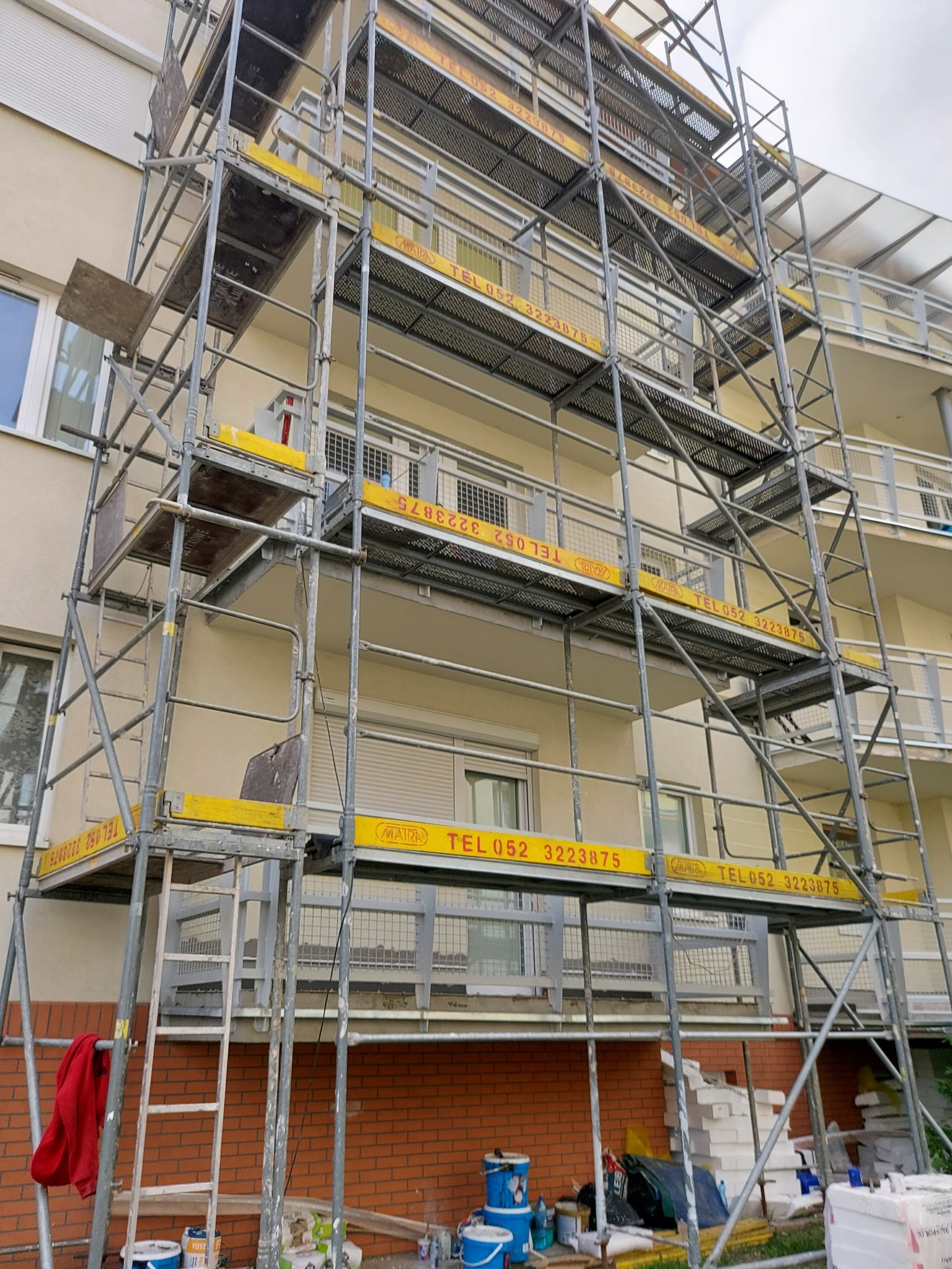 Budynek w rusztowaniach podczas prac remontowych. Widoczne farby i narzędzia malarskie. Ujęcie z dołu, ukazujące elewację w trakcie odświeżania.