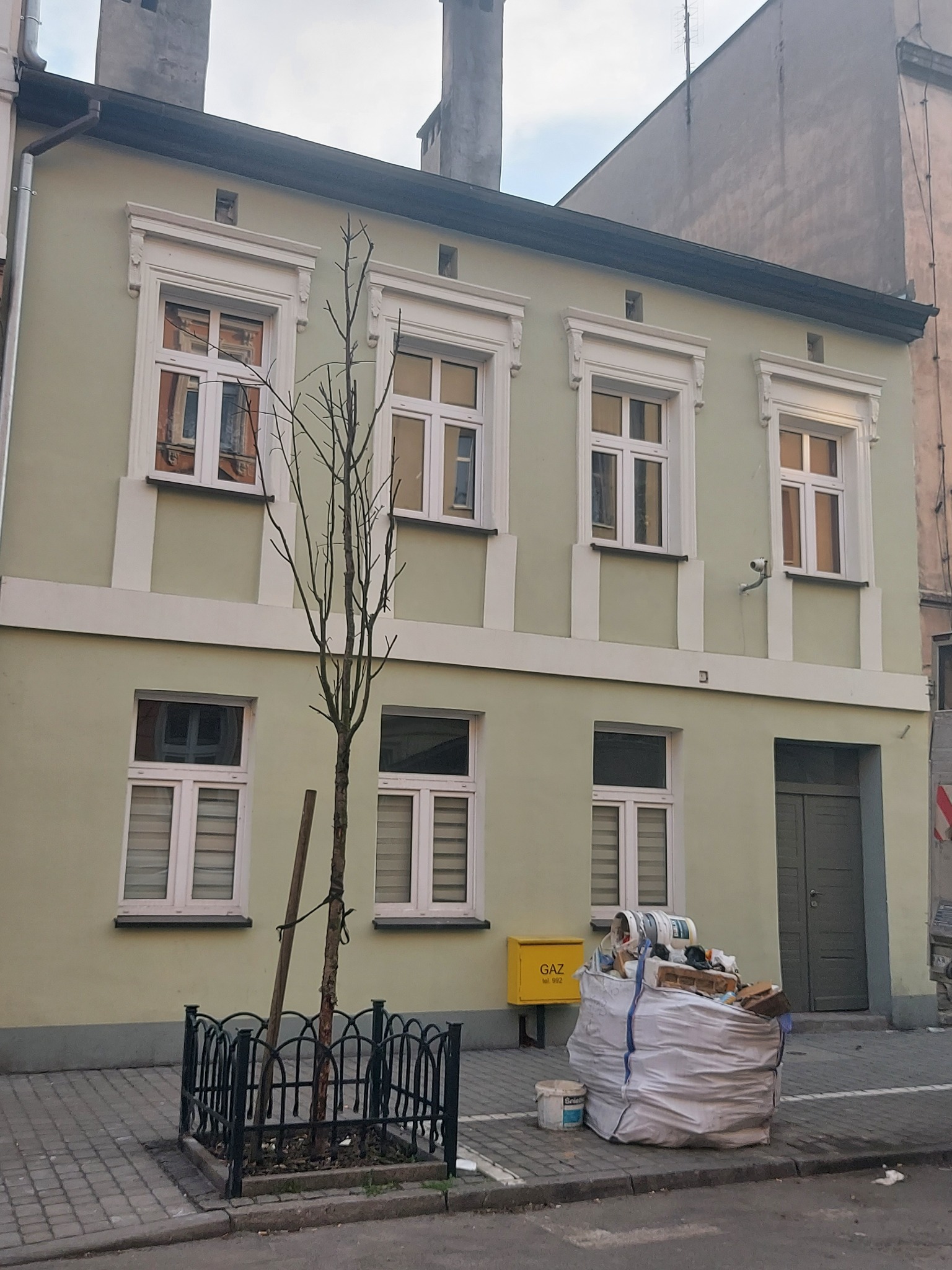 Odnowiona fasada kamienicy w pastelowej zieleni z białymi obramowaniami okien. U podnóża budynku widoczny worek z odpadami budowlanymi i skrzynka gazowa.