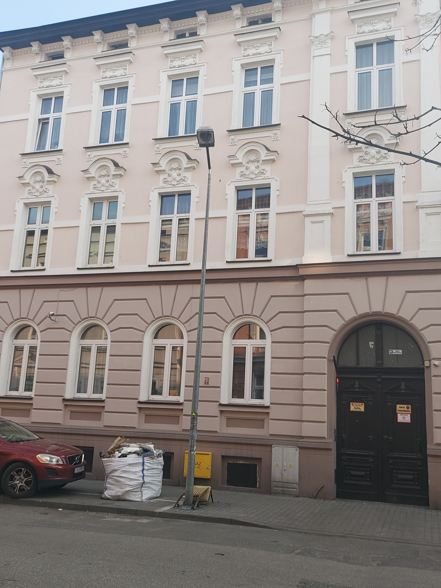 Odnowiona fasada kamienicy w pastelowych barwach z ozdobnymi oknami i nową stolarką. Na pierwszym planie worek z odpadami budowlanymi i zaparkowany samochód.