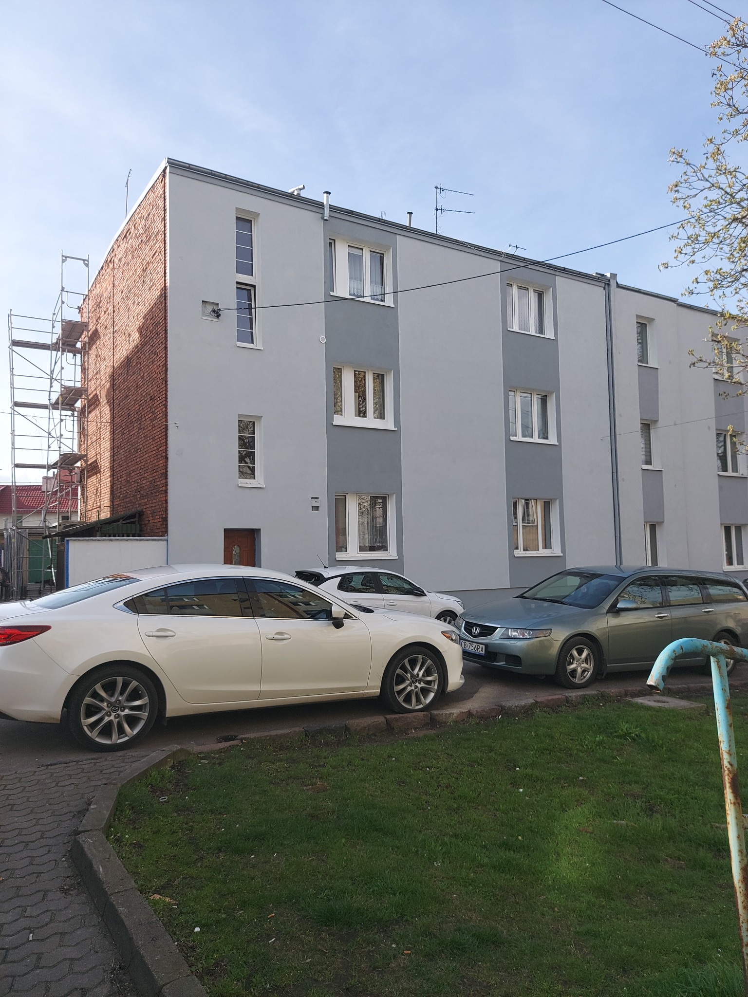 Odnowiona elewacja budynku wielorodzinnego w odcieniach szarości, z rusztowaniem przy ceglanej ścianie szczytowej. Zaparkowane samochody na pierwszym planie.