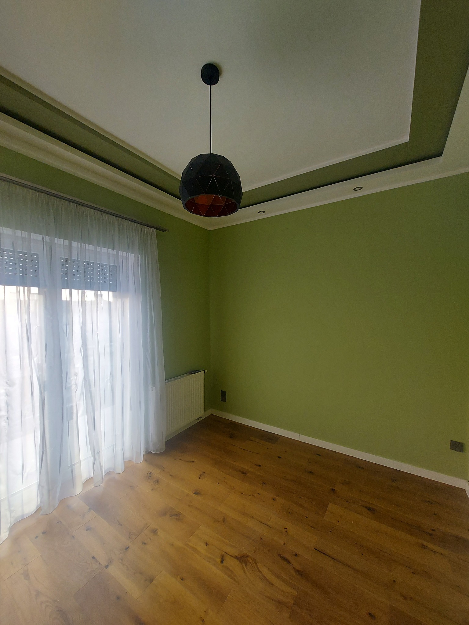 Pokój po malowaniu w Bydgoszczy: zielone ściany, drewniana podłoga, biały sufit z dekoracyjną listwą i nowoczesna lampa wisząca. Jasne zasłony w oknie.