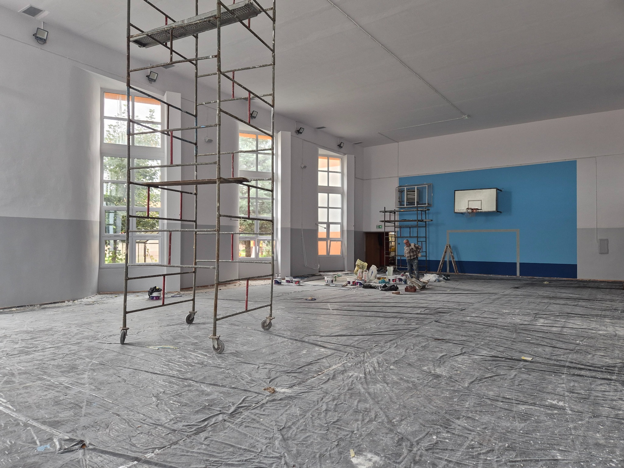 Malowanie sali gimnastycznej w trakcie prac. Widoczne rusztowania, zabezpieczona podłoga folią, puszki z farbą i malarz przy ścianie z koszem.