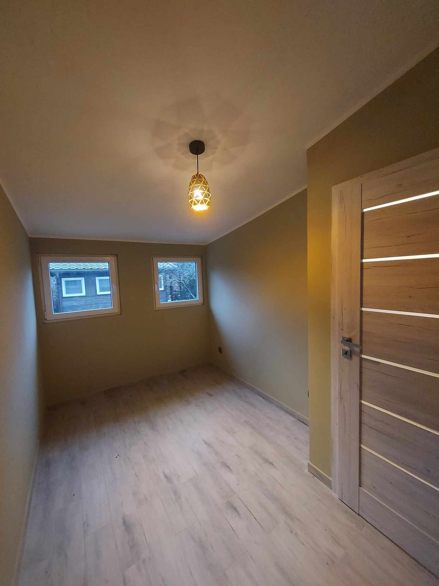 Pokój po remoncie: świeżo pomalowane ściany w odcieniach beżu, jasna podłoga imitująca drewno, nowoczesne drzwi z podświetleniem i designerska lampa.