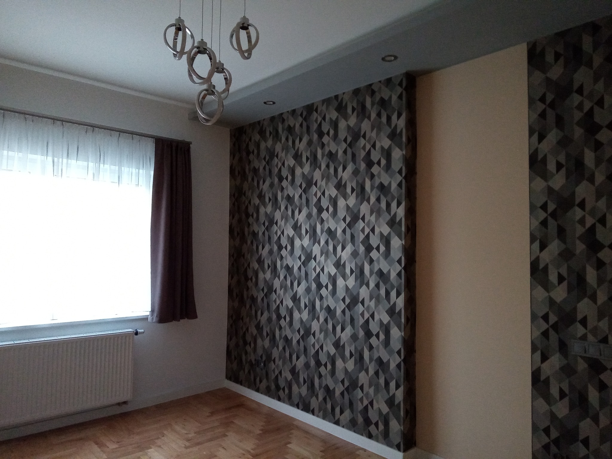 Pokój po tapetowaniu: ściana z geometrycznym wzorem w odcieniach szarości, brązowa zasłona przy oknie, drewniana podłoga i nowoczesna lampa sufitowa.